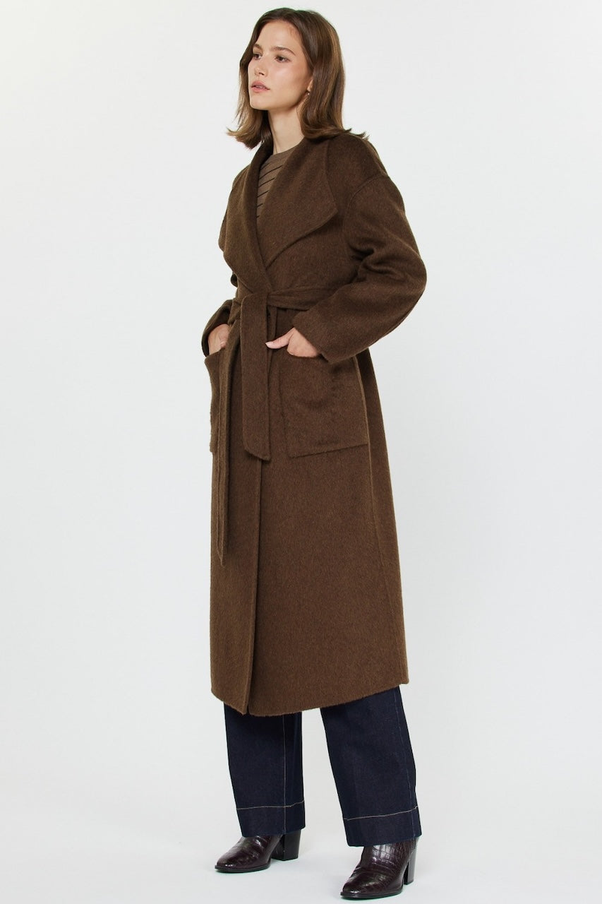 Woman wearing long brown wool coat with oversized fit and belted waist styled over jeans and boots.