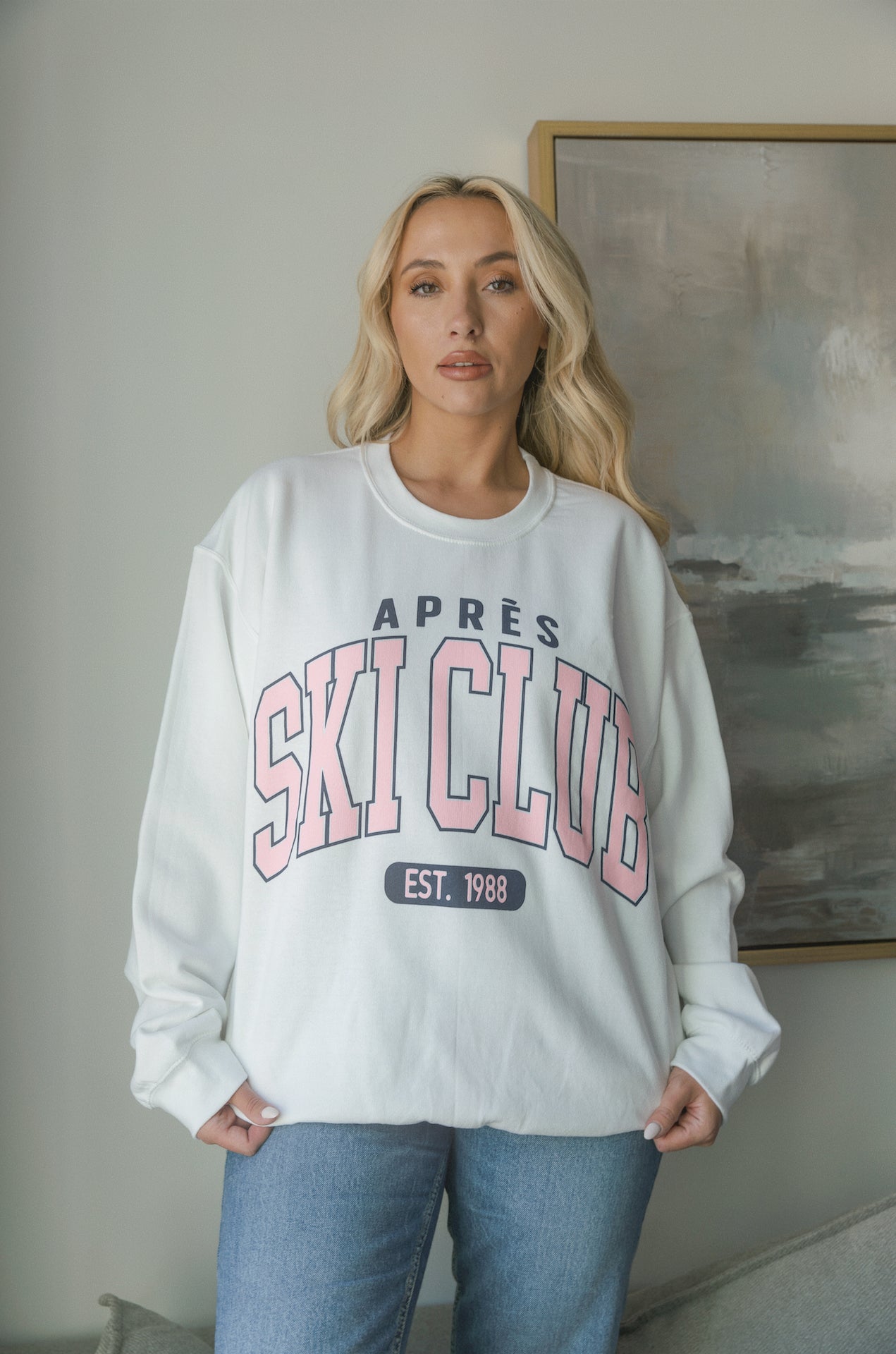 Model wearing oversized Apres Ski Club graphic sweatshirt in white with pink lettering, styled with denim jeans. Cozy apres sweatshirt with relaxed fit perfect for winter or ski trips.