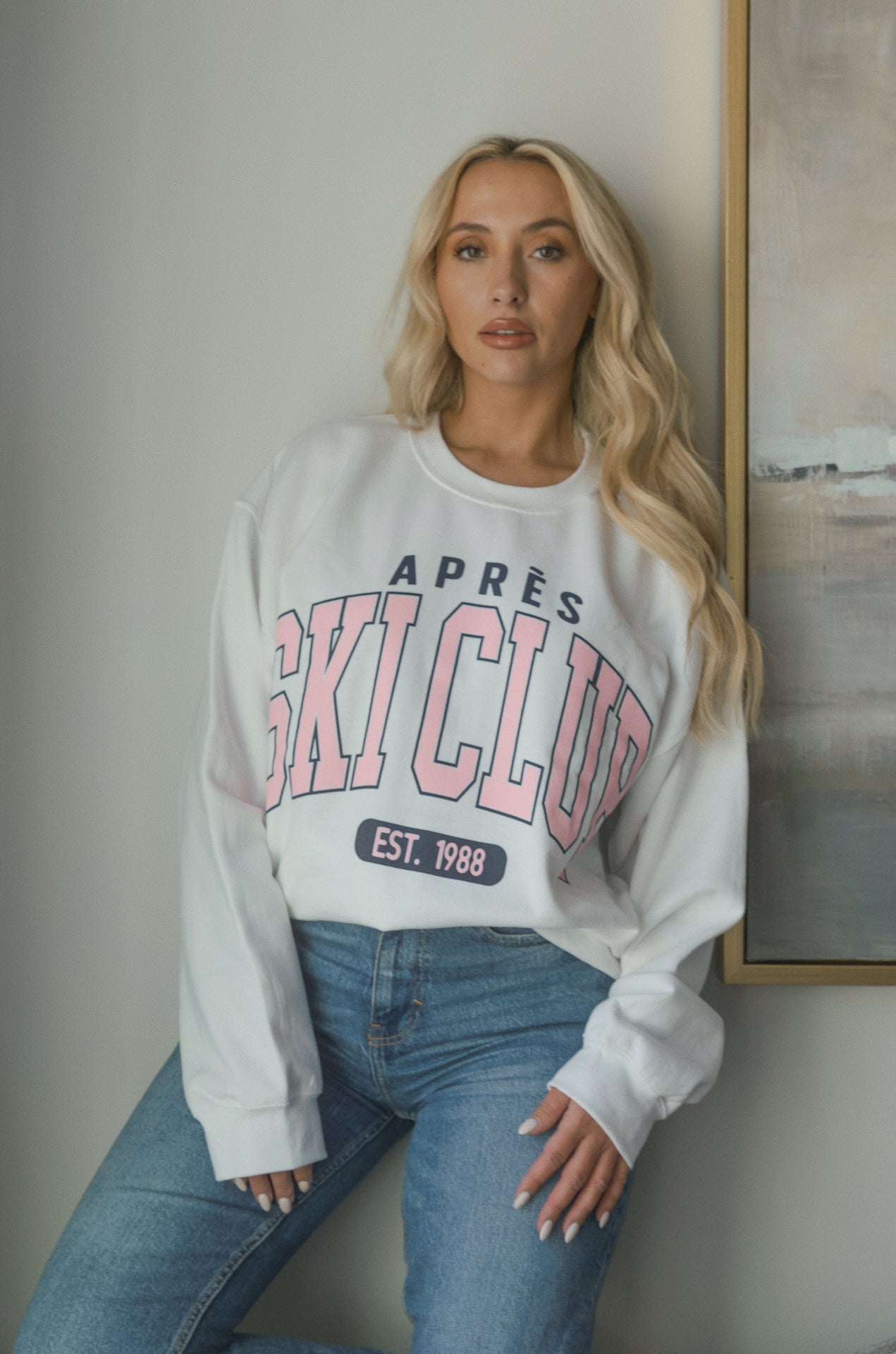 Model wearing oversized Apres Ski Club graphic sweatshirt in white with pink lettering, styled with denim jeans. Cozy apres sweatshirt with relaxed fit perfect for winter or ski trips.
