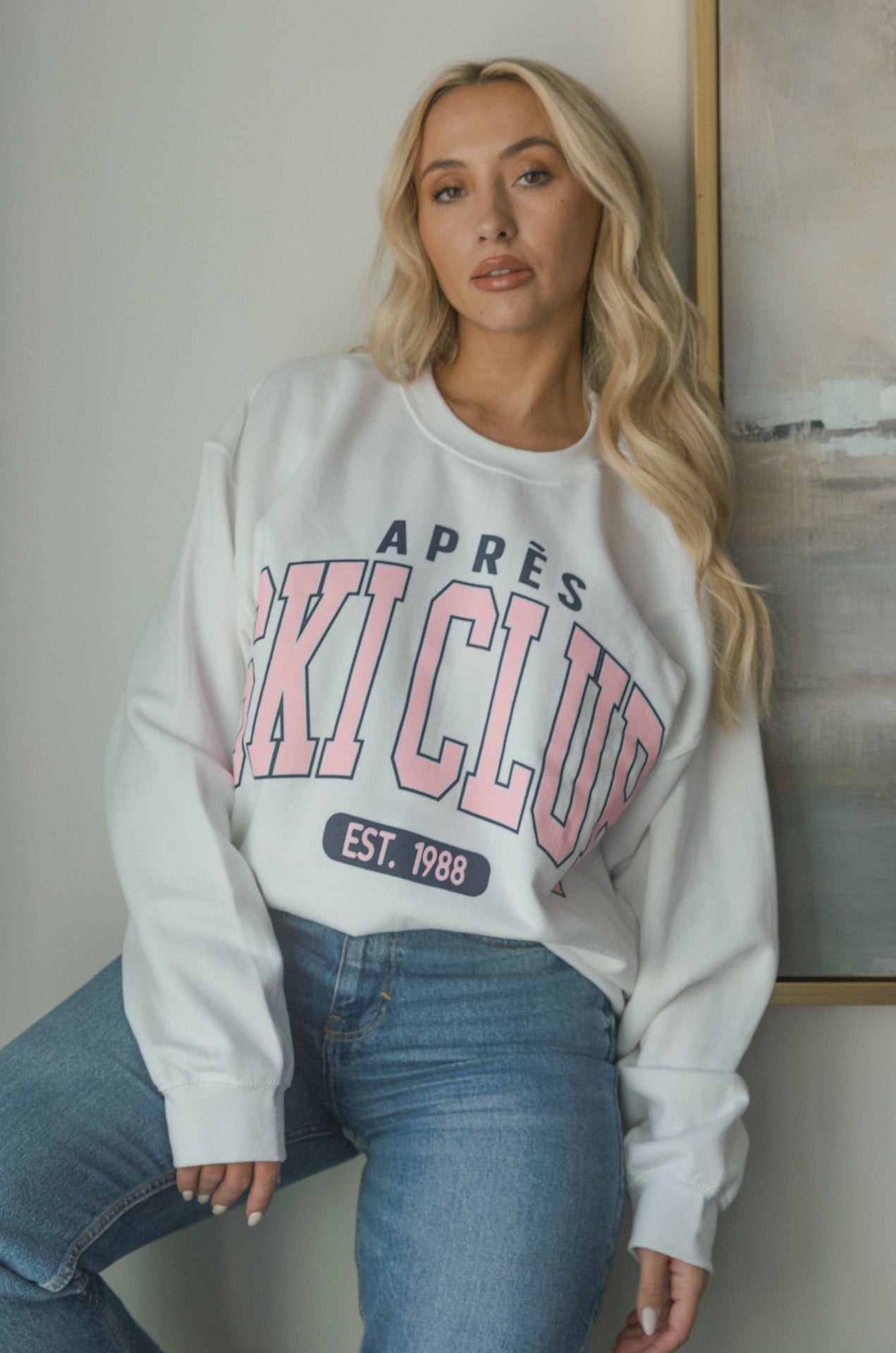 Model wearing oversized Apres Ski Club graphic sweatshirt in white with pink lettering, styled with denim jeans. Cozy apres sweatshirt with relaxed fit perfect for winter or ski trips.