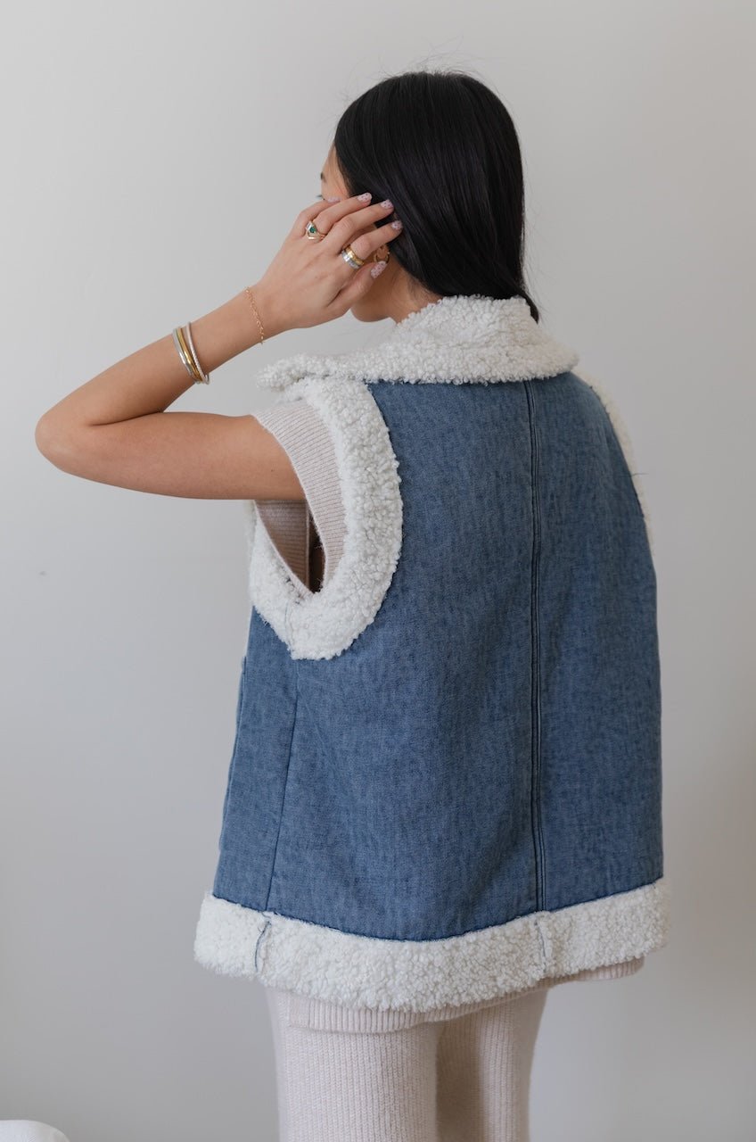 Person wearing a blue denim vest with white fur trim against a plain background