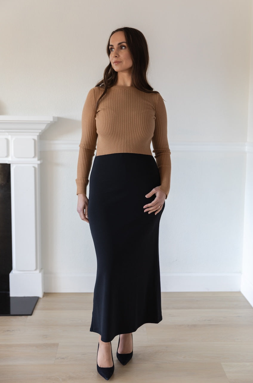 Woman wearing a fitted brown long sleeve crop top with ribbed texture and crew neckline, styled with a black fitted high waisted midi skirt.