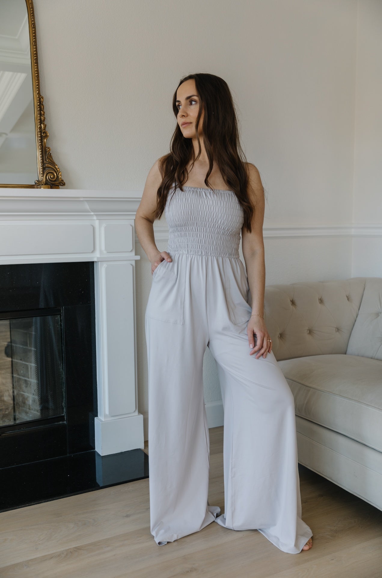 A woman standing in a room with a grey jumpsuit, sleeveless with a smocked bodice and wide-leg pants.