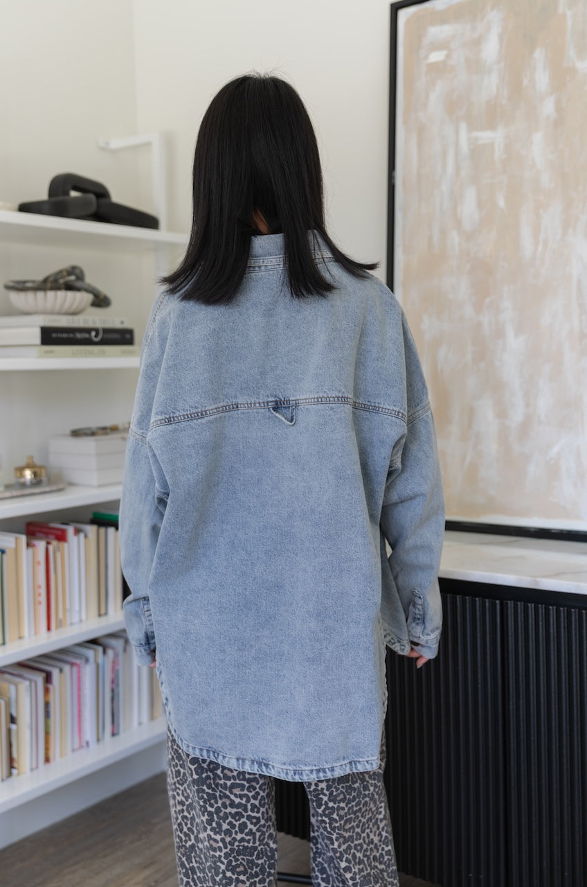 Woman wearing a light blue denim jacket in a room with a bookshelf and wall.