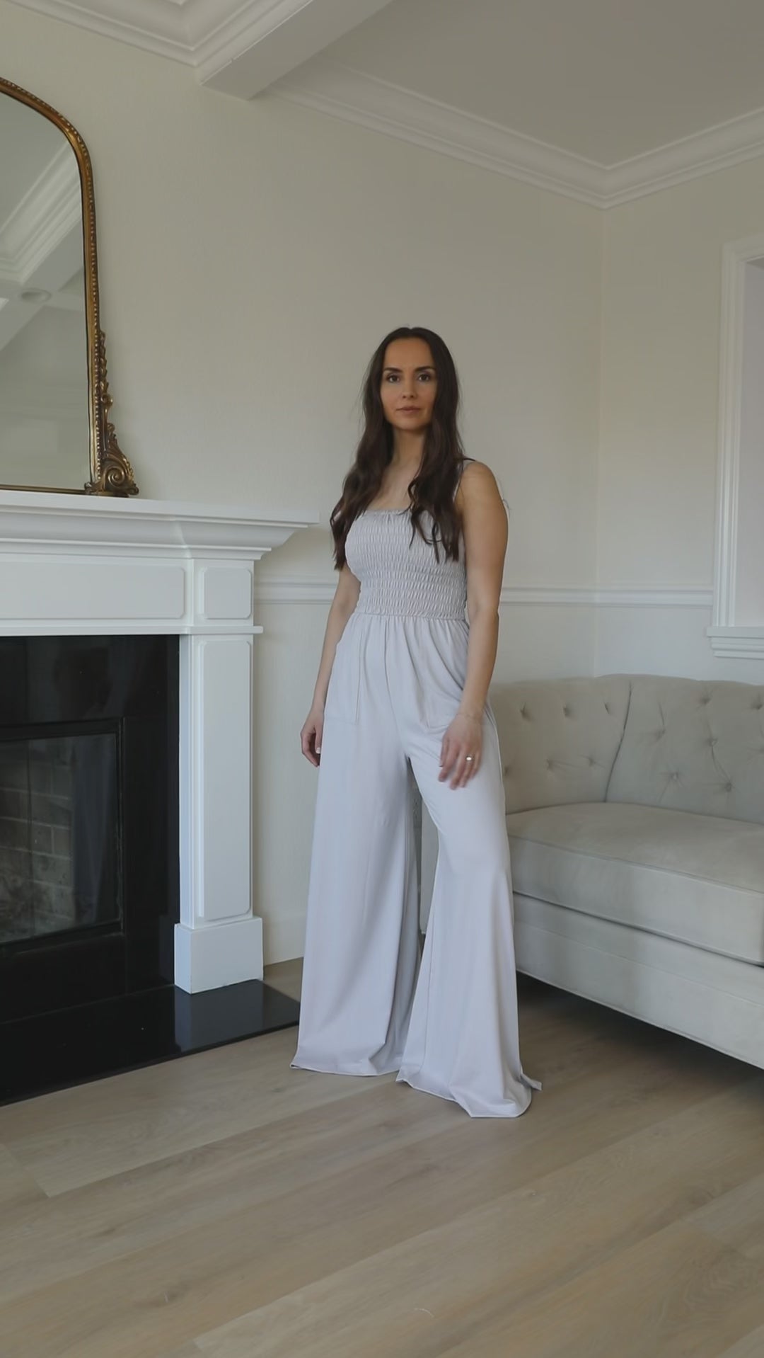 A woman standing in a room with a grey jumpsuit, sleeveless with a smocked bodice and wide-leg pants.