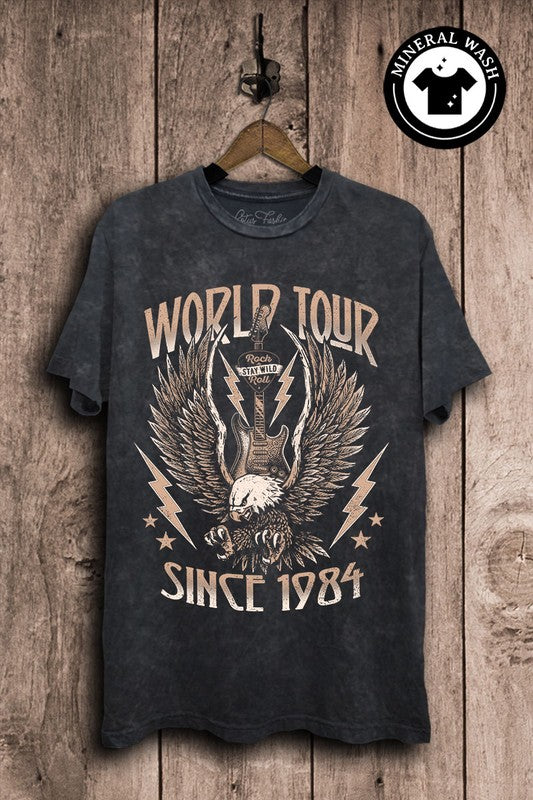 A black oversized graphic tee with 'WORLD TOUR SINCE 1984' printed on the front, displayed on a wooden hanger against a wood backdrop.
