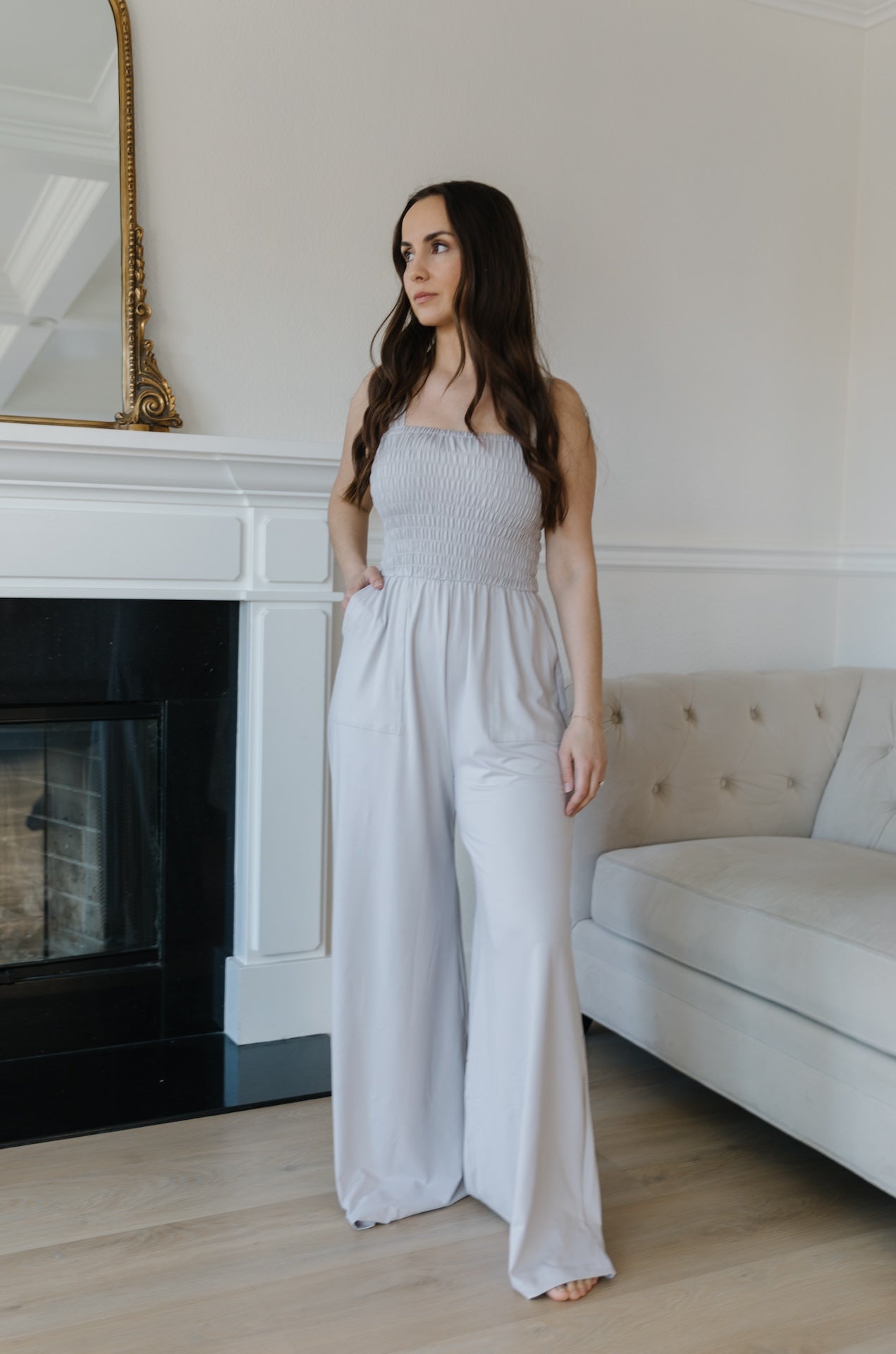 A woman standing in a room with a grey jumpsuit, sleeveless with a smocked bodice and wide-leg pants.