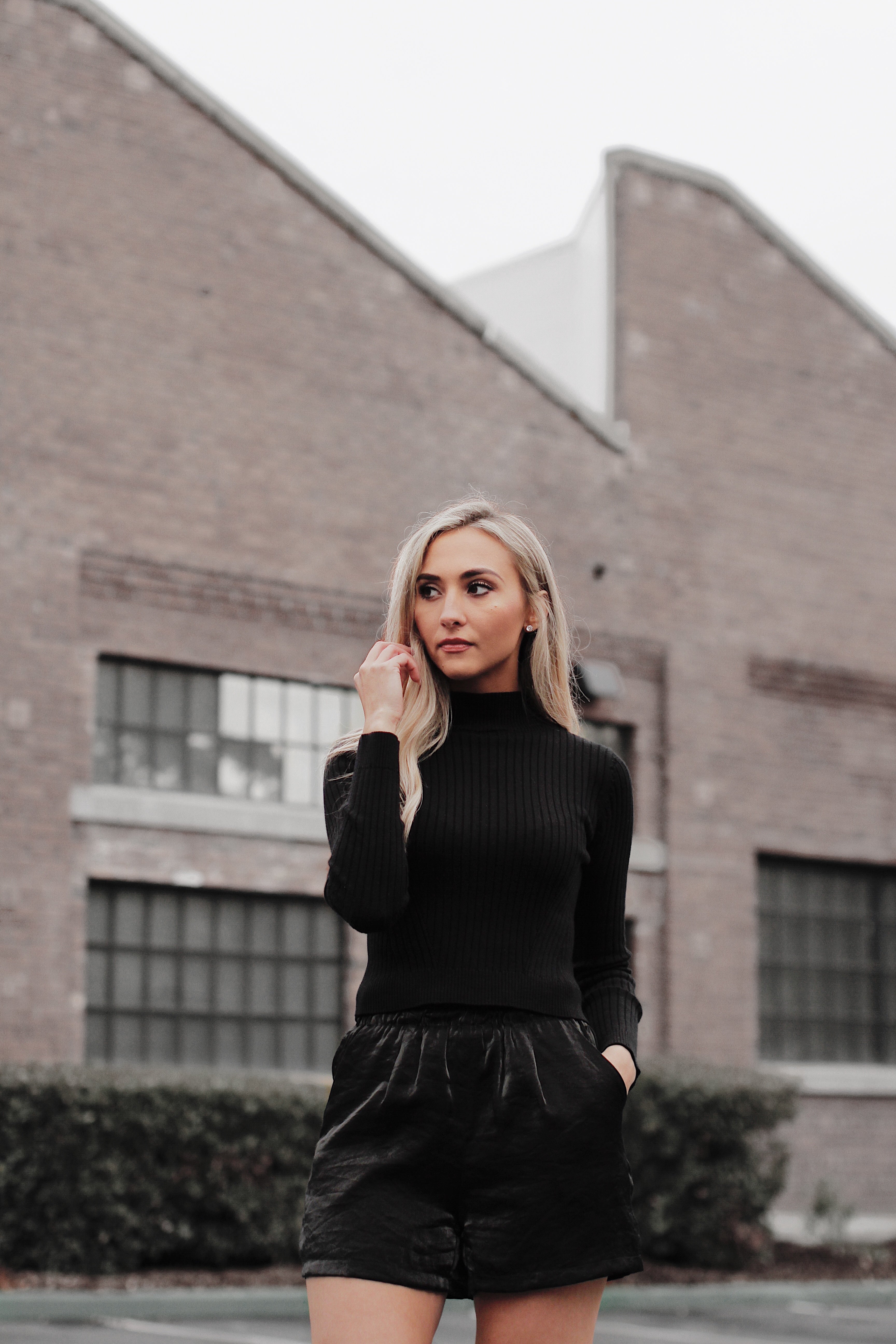 A woman wearing a black high waist formal shorts with side pockets and a black top, standing in front of a brick building.