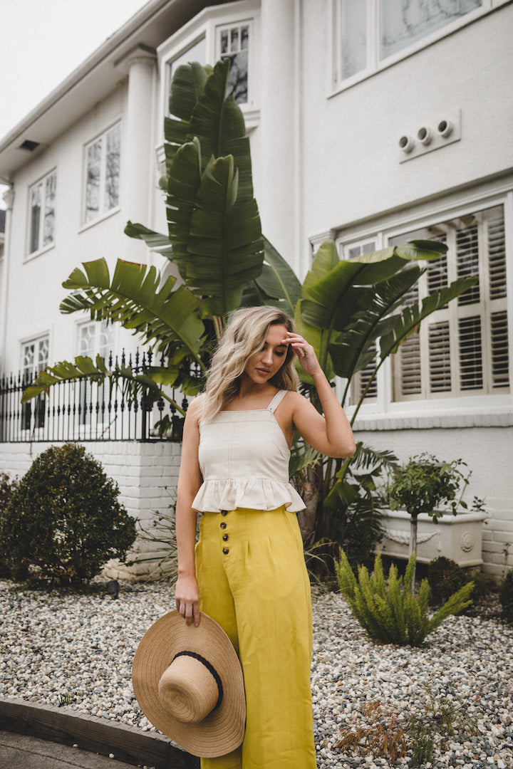 A woman wearing a beige linen sleeveless crop top with ruffle trim and adjustable straps, styled in a babydoll silhouette.
