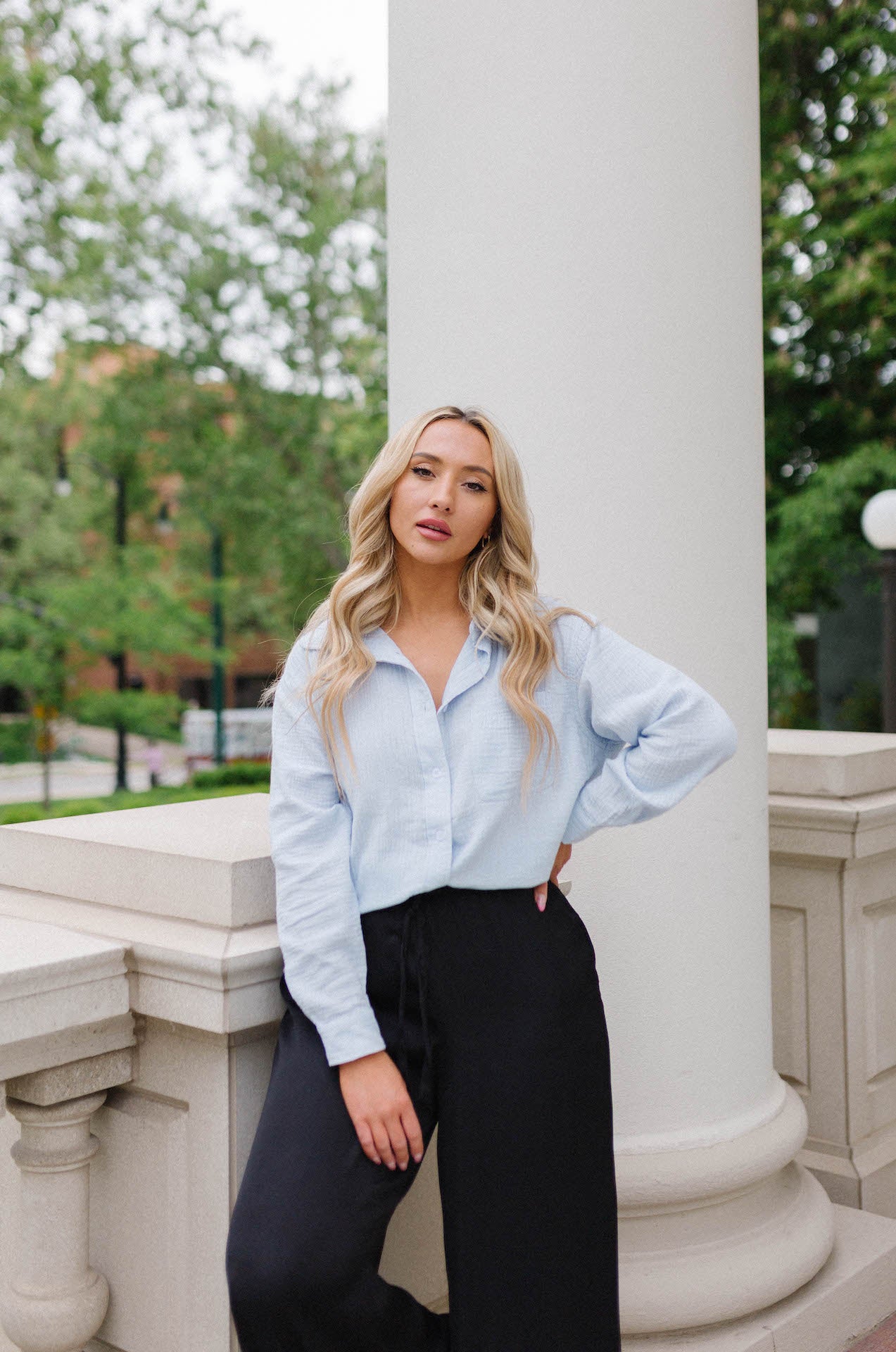 A woman wearing a blue long sleeve button-down shirt with a relaxed fit, standing near a column.